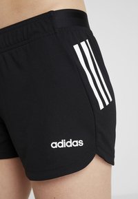 Primer plano de unos pantalones cortos deportivos negros de Adidas con el logo blanco y tres rayas blancas en el muslo izquierdo, usados por una persona.