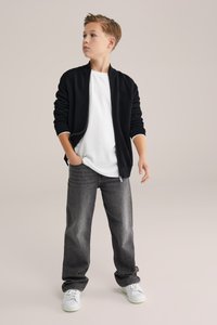 Svart jacka med dragkedja över en vit T-shirt, kombinerad med löst sittande grå jeans och vita sneakers. Enkel, vardaglig outfit.