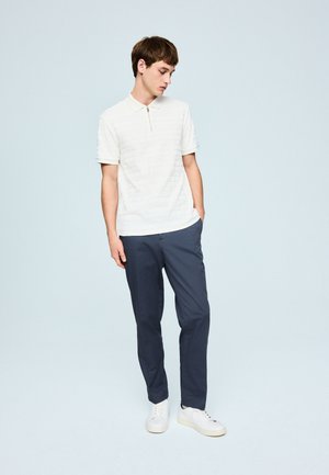 Ung mand iført hvid poloshirt med korte ærmer, mørkeblå bukser og hvide sneakers, står med den ene hånd i lommen.