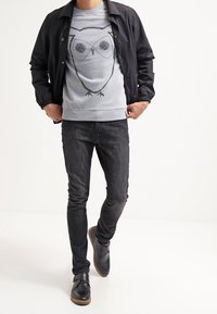 Czarna lekka kurtka na szarym swetrze z grafiką sowy, w połączeniu z ciemnymi jeansami i czarnymi butami wsuwanymi z brązowymi podeszwami.