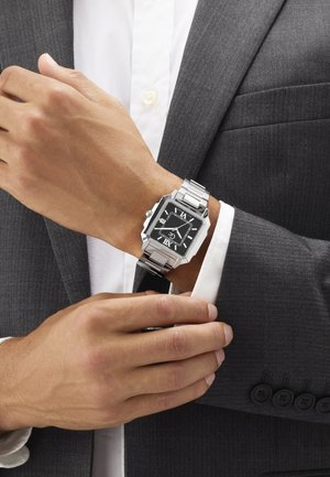 Polshorloge met een zwart vierkant horlogeplaat, Romeinse cijfers en een zilveren metalen band, gedragen aan de linkerpols en versteld met de rechterhand over een grijze mouw van een pak.