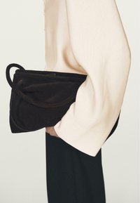 Braune Wildleder-Handtasche mit geschwungenem Design und einem gewellten Griff. Die Tasche wird vor einem hellfarbenen Kleidungsstück und einem schwarzen Unterteil gehalten.