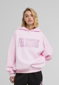 Růžová mikina s textem "FELICIOUS" ve třpytivém designu. Má velkou kapuci, oversize rukávy a měkkou látku. V kombinaci s černými kalhotami.