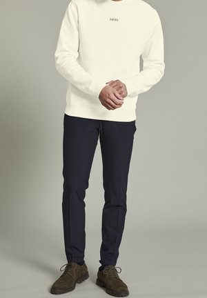 Mies, joka pukeutunut valkoiseen collegepaitaan, tummansinisiin slim-fit-housuihin ja ruskeisiin nauhallisiin kenkiin, seisoo yksinkertaisen taustan edessä.