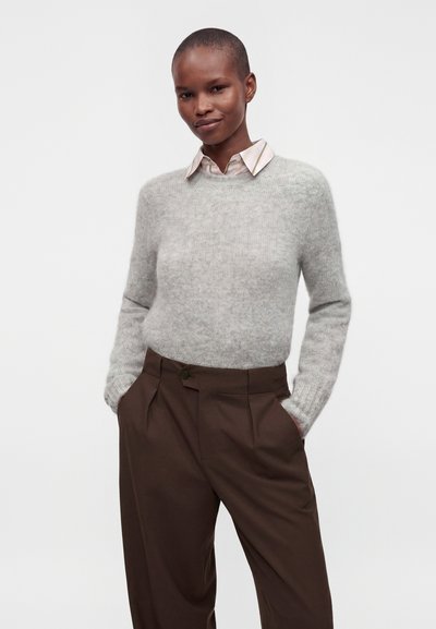 Personne debout, les mains dans les poches, portant un pull gris clair sur une chemise à col et un pantalon marron taille haute, fond neutre.