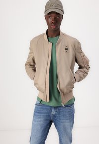 Beige bomberjakke med ribbestrikkede mansjetter, glidelås og logodetalj, kombinert med en grønn t-skjorte og blå jeans, fullført med en utslitt caps.
