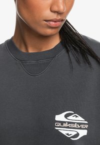 Grå genser med ribbestrikket hals og mansjetter, med en hvit Quiksilver-logo på venstre side, laget av mykt stoff med en teksturert overflate.