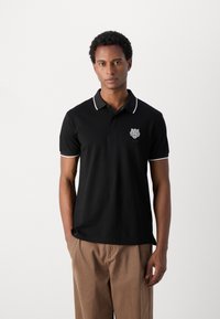 Zwarte polo met witte kraag en accenten op de mouwen, voorzien van een geborduurd leeuwenlogo op de borst. Draagt dit met beige broek.