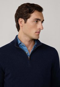 Mann mit kurzen braunen Haaren, der einen dunkelblauen Half-Zip-Pullover über einem blauen Button-Up-Hemd trägt und nach rechts schaut, vor einem einfarbigen Hintergrund.