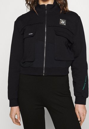 Kobieta ubrana w czarną kurtkę na suwak z dwoma przednimi kieszeniami na klapy i logo "Puma" na piersi i rękawie z napisem "Forever Better."