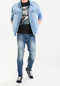 Ljusblå denimjacka med slitna detaljer, svart grafisk t-shirt, slim fit blå jeans och svarta sneakers med vita sulor.