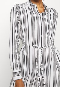 Abito a camicia a righe in tessuto bianco e nero. Maniche lunghe, cintura da annodare in vita e tasca sul lato sinistro del petto. Le righe verticali variano in larghezza.
