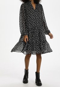 Svart veckad klänning med V-ringning, långa genomskinliga ärmar, vitt blommönster och en lagrad kjol; tillsammans med svarta ankelstövlar.