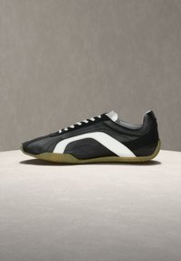 Μαύρο αθλητικό παπούτσι χαμηλού τύπου με λευκή πλευρική ρίγα και κορδόνια, καφέ υφασμένο πάτο, τοποθετημένο σε φωτεινή επιφάνεια με γκρι φόντο.