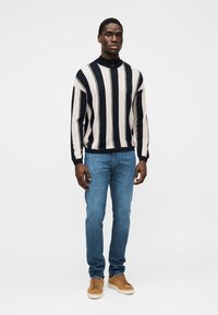 Črno-krem pulover z visokim ovratnikom, kombiniran s svetlo modrimi kavbojkami in svetlo rjavimi supergami. Enostavna, oprijeta zasnova s kontrastnimi barvami.