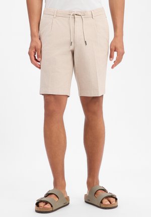 Pantaloncini beige al ginocchio con pieghe frontali e vita con coulisse indossati da una persona in piedi con pelle abbronzata e sandali.