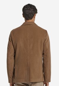 Braune Cordjacke mit Kragen, strukturiertem Stoff und klarer Form. Sichtbare Nahtdetails und gerader Schnitt am Saum.