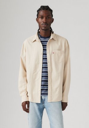 Camisa de botones beige de manga larga, con un bolsillo delantero, llevada sobre una camiseta de rayas azul marino, combinada con jeans de mezclilla azul claro.