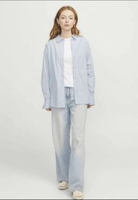 Světle modrá a bílá pruhovaná dlouhá košile, nošená přes bílý tričko, v kombinaci se světlými širokými džíny. Texturované pletené boty.