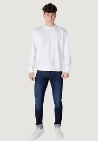 Felpa bianca a maniche lunghe con scollatura tonda, abbinata a jeans blu scuro e sneakers bianche. Design semplice, vestibilità classica.