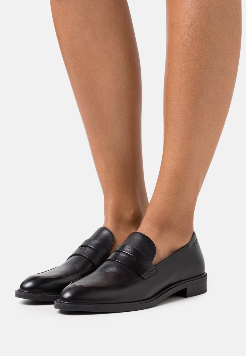 Mocassins en cuir noir avec un petit talon portés pieds nus, vus de face et de côté sur un fond uni.