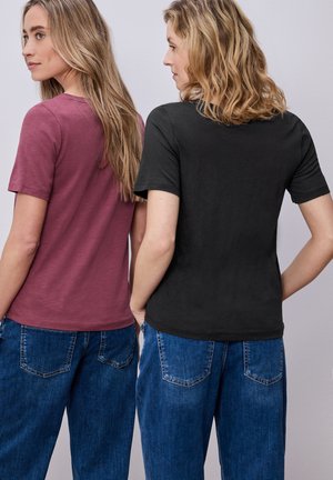Zwei Frauen stehen Rücken an Rücken, tragen lässige T-Shirts, eines in Burgunderrot und eines in Schwarz, kombiniert mit blauen Jeans.