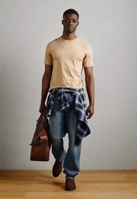 Camiseta de manga corta color beige, jeans azules, camisa de cuadros atada a la cintura, bolso de cuero marrón, zapatos marrones y fondo de suelo de madera.