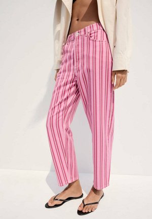 Mujer que lleva pantalones rosas de talle alto con rayas verticales, chanclas negras y una chaqueta corta beige, de pie frente a un fondo blanco.