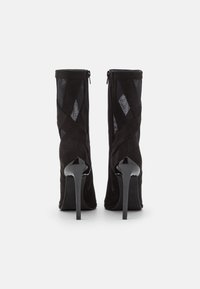 Bottines en daim noir et maille avec des talons aiguilles hauts et brillants, vues de dos sur un fond blanc.