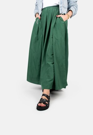 Green, pleated maxi skirt with an elastic waistband, front pockets, and black strappy sandals with an open toe and thick sole.