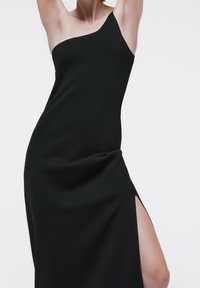 Abito nero monospalla con una silhouette slim e aderente. Presenta una fessura laterale, una texture liscia e dettagli drappeggiati sulla gonna.