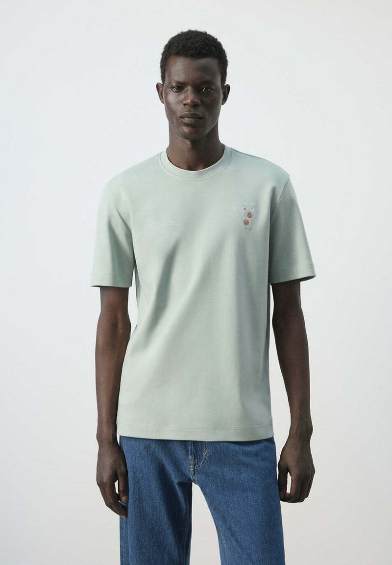 Homme portant un t-shirt manches courtes vert clair avec un petit motif floral et un jean bleu, debout devant un fond blanc uni.