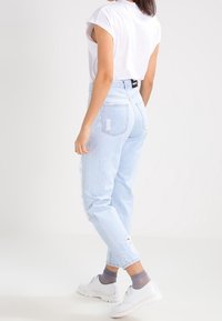 Jean denim bleu clair, usé, avec une coupe décontractée. Associé à une chemise blanche à manches courtes et des chaussures blanches. Chaussettes grises texturées visibles.