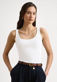 Biały prążkowany top na ramiączkach z zaokrąglonym dekoltem, zestawiony z ciemnymi spodniami i brązowym skórzanym paskiem z złotą klamrą i ozdobnym zawieszką.