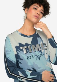 Maglietta a maniche lunghe di colore azzurro chiaro con motivo camo blu scuro, caratterizzata da una scritta argento "TIME to inspire" e dettagli a righe sulle maniche.