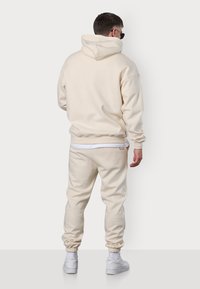 Cremefarbene Kapuzenjacke und Jogginghose aus weichem Stoff. Zu den Merkmalen gehören eine Kapuze, gerippte Bündchen und ein elastischer Bund; kombiniert mit weißen Turnschuhen.