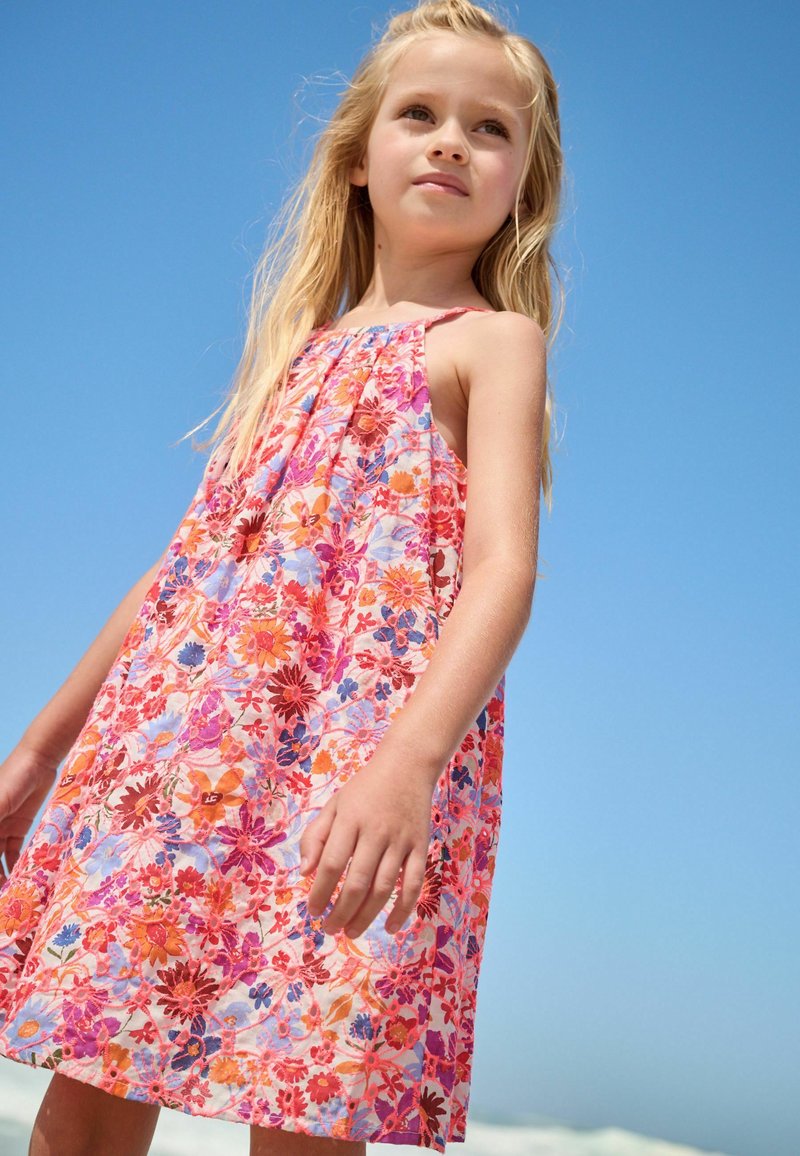 Jeune fille portant une robe fleurie colorée, debout à l'extérieur sous un ciel bleu clair.