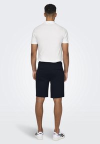 Hombre de pie de espaldas, vistiendo una polo blanca de manga corta, pantalones cortos negros con bolsillos traseros y zapatillas blancas con detalles negros.