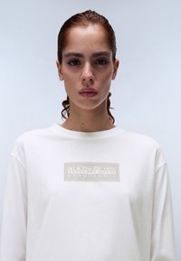 Valkoinen pitkähihaisen paita pehmeästä kankaasta, jossa on edessä vaaleanharmaa suorakulmainen logo, jossa lukee "NAPAPIJRI geographic".