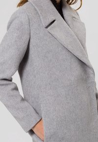 Cappotto grigio in misto lana con colletto a risvolto, maniche lunghe e tasche laterali. Il tessuto ha una finitura soffice e testurizzata.