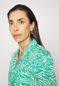 Blusa verde com padrão de zebra branco, design com colarinho, tecido leve. Brincos de argola dourados e colares em camadas delicados visíveis.