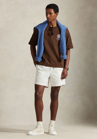 Καφέ κοντομάνικο T-shirt με λογότυπο, λευκές σορτς, μπλε πλεκτό γιλέκο περασμένο στους ώμους, μπεζ αθλητικά παπούτσια και ανοιχτόχρωμες κάλτσες.