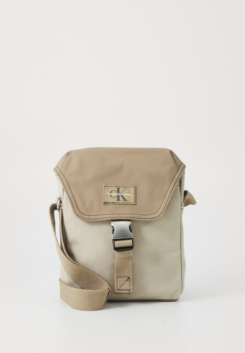 Svetlohnedá plátenná crossbody taška s klopou, striebornými kovovými doplnkami a logom. Má textilný popruh na nosenie.