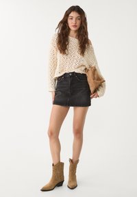 Pull en laine crème à mailles ouvertes, associé à une mini-jupe en denim noir, des bottines en daim marron, et un sac à main assorti.