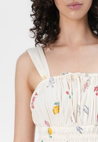 Vrouw met krullend haar in een witte mouwloze jurk met kleurrijke fruit- en bloemafdrukken, gestrikt op de borst met een strik.