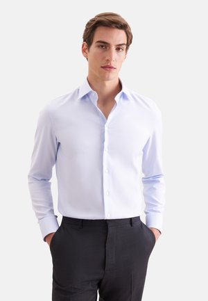 Jeune homme portant une chemise bleu clair à col ouvert et un pantalon habillé sombre, debout les mains dans les poches sur fond blanc.