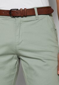 Lichtgroene broek van een gladde stof, met een bruine leren riem met een metalen gesp, vastgemaakt via lusjes in de taille.