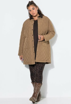 Mujer que lleva un abrigo acolchado marrón claro, leggings con estampado de leopardo y botas vaqueras marrones, de pie frente a una pared blanca lisa.