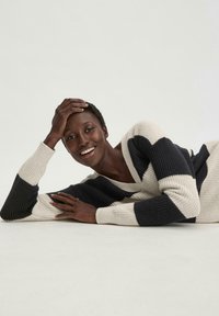 Strickpullover mit großen horizontalen Streifen in Marineblau und Creme. Verfügt über einen V-Ausschnitt und lange Ärmel mit Rippbündchen, gezeigt an einem Modell, das liegend dargestellt wird.