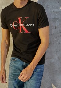 Svart bomullströja med en stor röd och vit "Calvin Klein Jeans"-logotyp. Korta ärmar och en klassisk passform, kombinerad med blå jeans.